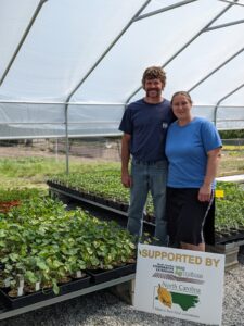 Two people in a green house stand behind a Supported by North Carolina sign.