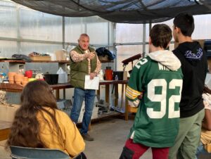 Students ask a man in a greenhouse questions.