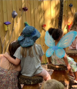 Children wearing colorful wings sit outside and enjoy story time. A young girl is hugging another young girl while they listen.