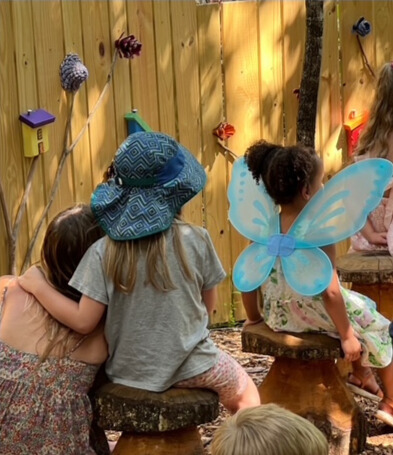 Children wearing colorful wings sit outside and enjoy story time. A young girl is hugging another young girl while they listen. 