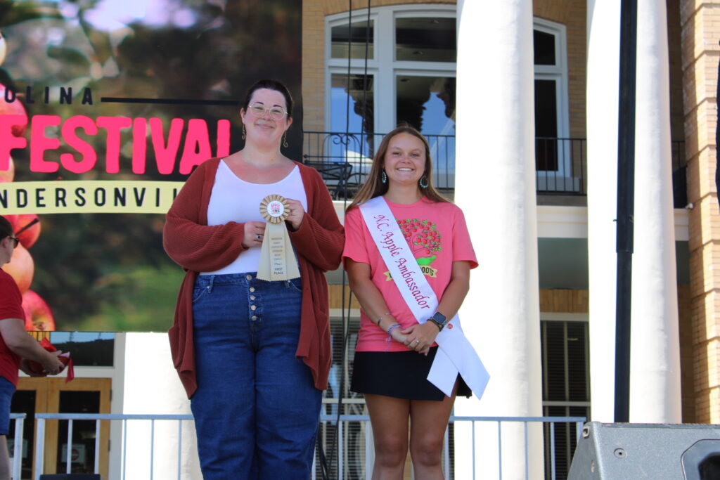  A full-length photo shows two women on an outdoor stage. The woman on the left is wearing a white top and a rust-colored cardigan, and she is holding a "Breads 1st Place" ribbon. The woman on the right is wearing a pink T-shirt and a white "NC Apple Ambassador" sash. Both are smiling at the camera. A large screen behind them reads "North Carolina Apple Festival, Hendersonville."