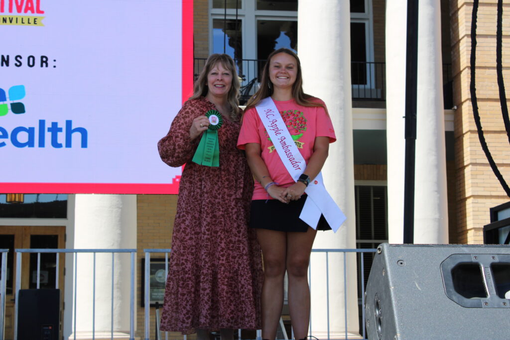 A woman with a long floral dress and a "Breads 3rd Place" ribbon is standing next to a young woman wearing a pink T-shirt and an "NC Apple Ambassador" sash. They are on an outdoor stage, smiling for the camera. A large screen behind them reads "Apple Festival."