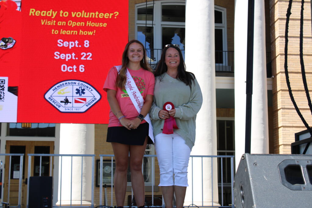 Two women are standing on an outdoor stage. The  woman on the left is wearing a pink T-shirt, a black skirt, and a white "NC Apple Ambassador" sash. The woman on the right is wearing a green sweater and white pants, and she is holding a red ribbon that says "Desserts 2nd Place." Both are smiling at the camera. A large red banner is behind them.