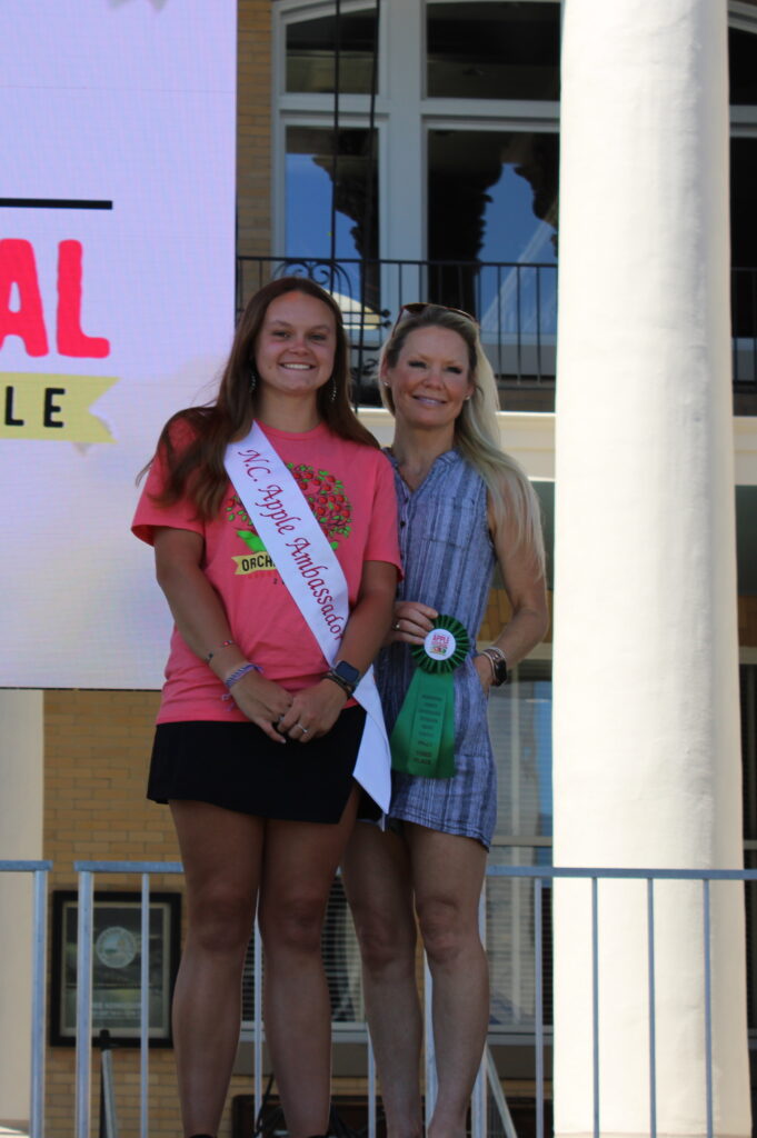 A photo shows two women on an outdoor stage. The woman on the left is wearing a pink T-shirt and a white sash that says "NC Apple Ambassador." The woman on the right is wearing a blue patterned dress and holding a green ribbon. Both are smiling at the camera. In the background, a large screen reads "North Carolina Apple Festival, Hendersonville."
