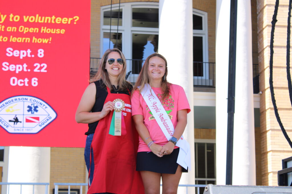 Two women stand on an outdoor stage. The woman on the left wears a red apron and holds a "Grand Champion" ribbon. The woman on the right wears a pink shirt and a sash that says "NC Apple Ambassador." Both are smiling at the camera.