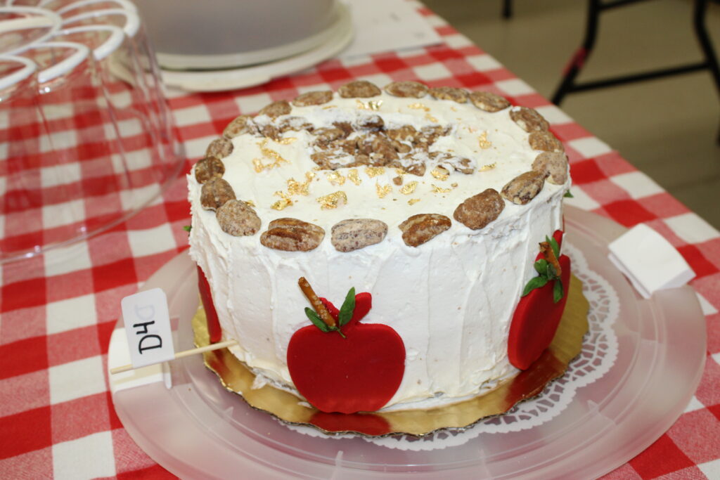 A frosted cake sits on a table with a red and white checkered tablecloth. The cake is decorated with red fondant apples, pecans around the top edge, and what appears to be gold leaf flakes. A small white flag with "D4" written on it is stuck into the side of the cake.