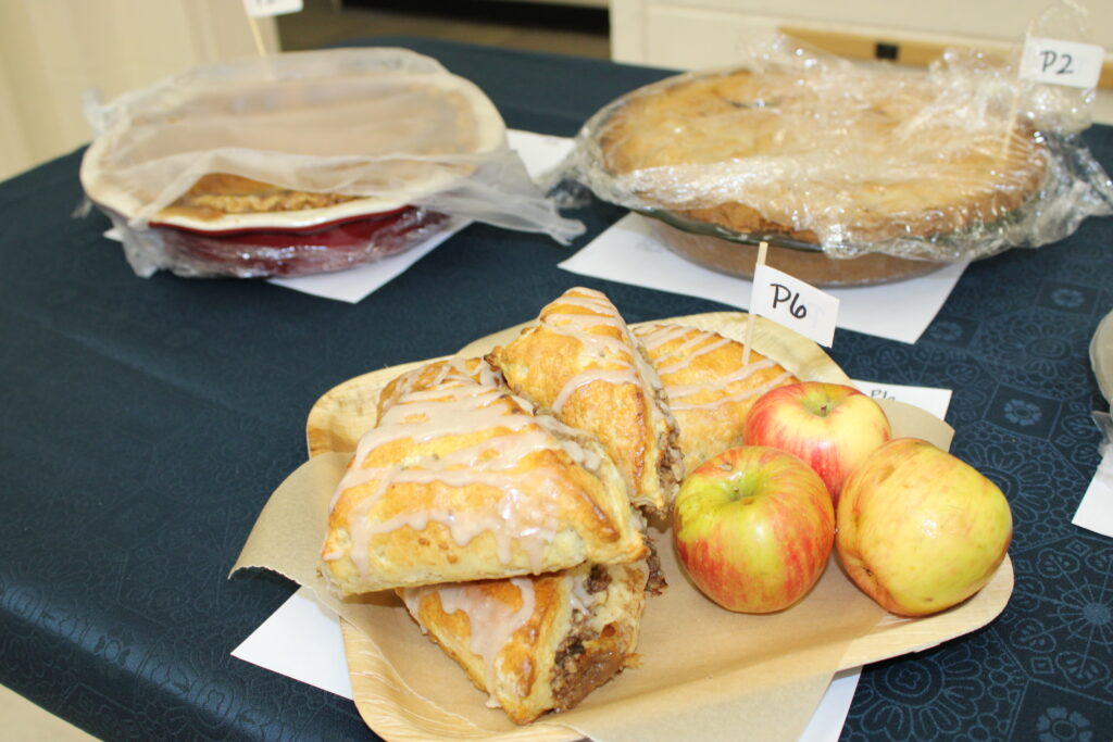  A light-colored plate holds three apple turnovers drizzled with white icing, along with three small apples. A small white flag with "P6" written on it is stuck into one of the turnovers.
