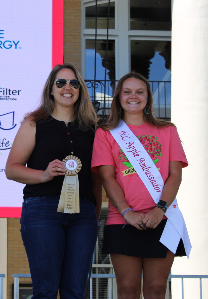  A woman in a black top and a woman in a pink T-shirt stand on a stage, smiling. The woman on the left is holding a ribbon, while the woman on the right is wearing a sash that says "N.C. Apple Ambassador."
