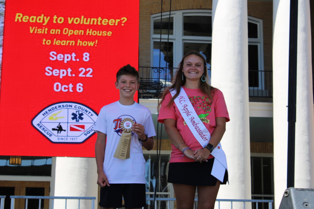  A smiling boy in a white T-shirt and black shorts stands next to a young woman wearing a pink T-shirt, black skirt, and a white sash that says "NC Apple Ambassador." The boy is holding a "Kids 1st Place" ribbon. They are on an outdoor stage with a large red banner behind them.