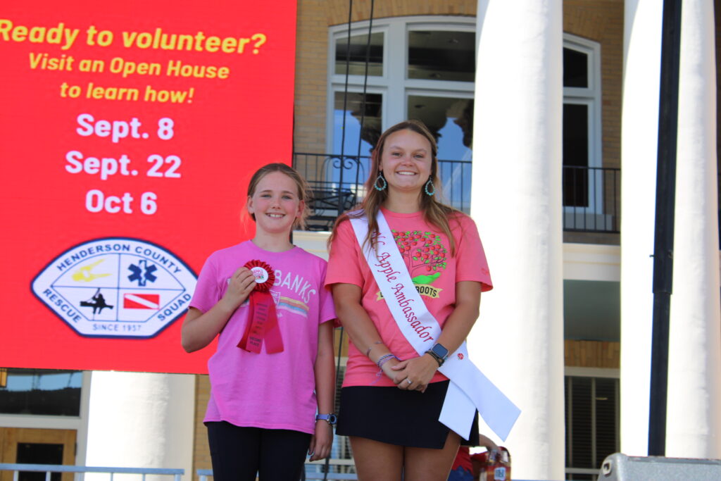 A girl and a young woman are standing on an outdoor stage. The girl on the left, wearing a pink T-shirt, holds a red ribbon that says "Kids 2nd." The young woman on the right is also wearing a pink T-shirt, a black skirt, and a white sash that says "NC Apple Ambassador." Both are smiling at the camera. A large red banner is behind them.