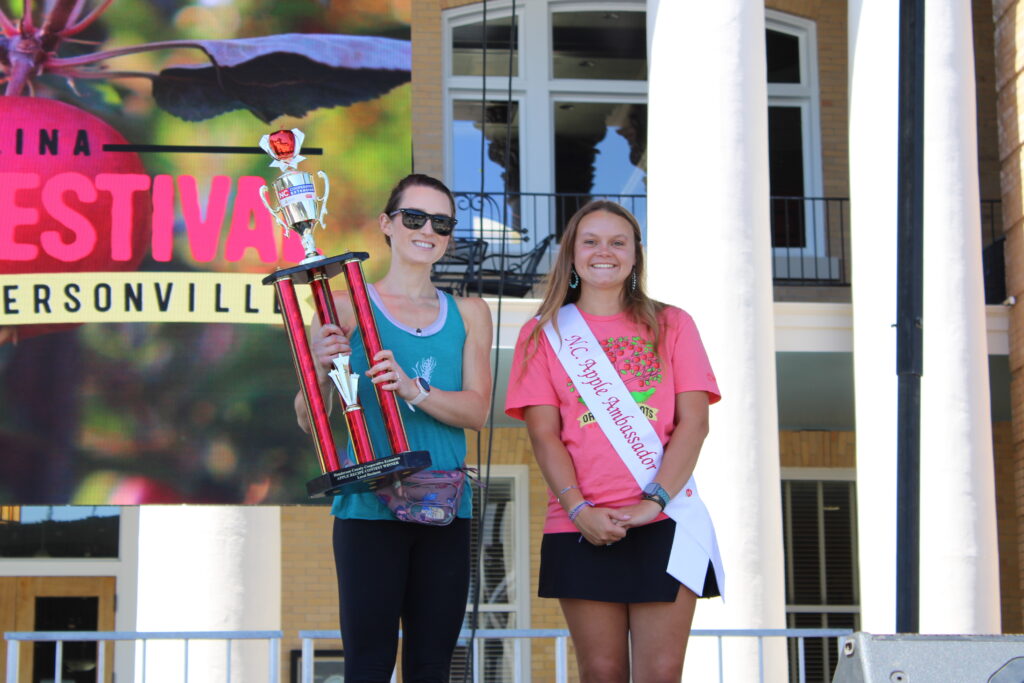A smiling woman in a teal tank top is standing on an outdoor stage, holding a large trophy. Next to her is a smiling young woman wearing a pink T-shirt and a white sash that says "NC Apple Ambassador." A large screen behind them reads "North Carolina Apple Festival, Hendersonville."
