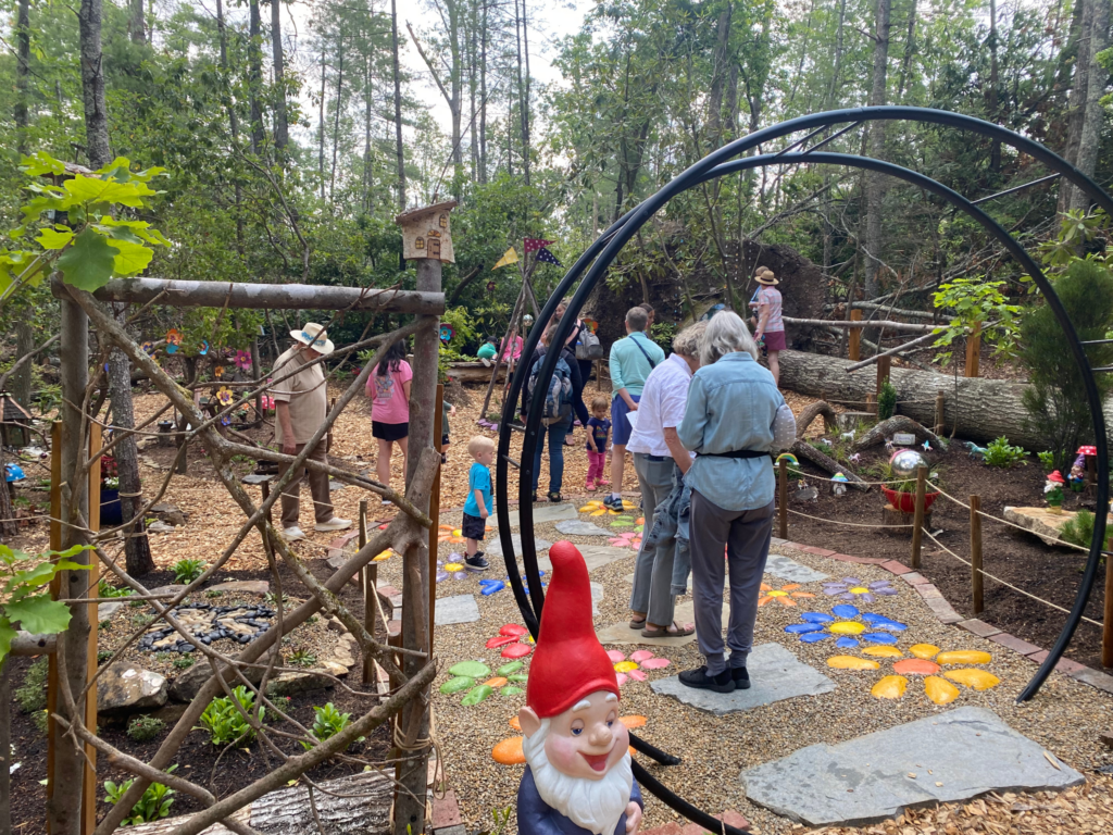 Adults and children walk on an outdoor trail with fairies, gnomes, and flowers.
