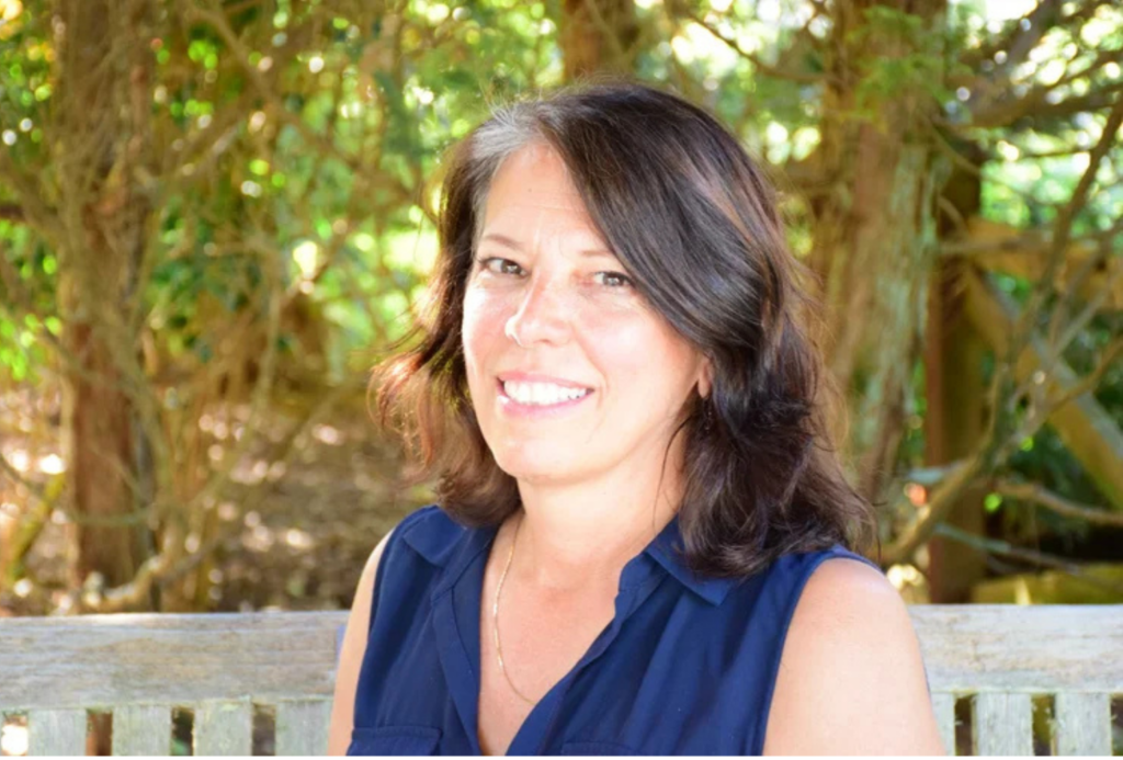 A smiling woman with dark hair and gray streaks sits on a wooden bench outdoors, wearing a navy blue sleeveless collared shirt. The background is filled with green foliage and trees.