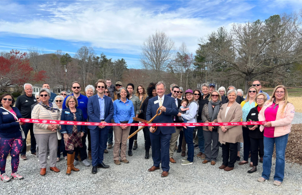 A diverse group of around 40 people stands outside on a gravel path, smiling for a photo behind a red ribbon. A man in a dark suit in the center holds a large pair of scissors, ready for a ribbon-cutting ceremony. Trees and hills are in the background under a blue sky.