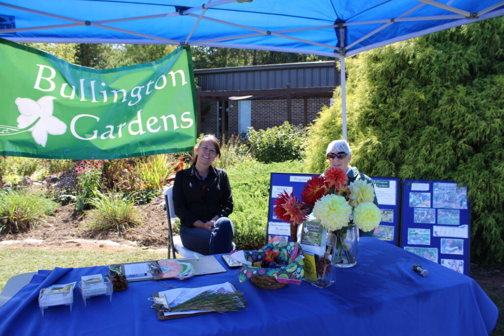 Two women sit at a Bullington Gardens information table under a blue canopy outdoors. The table is covered in a blue cloth and displays flowers, brochures, and informational posters.