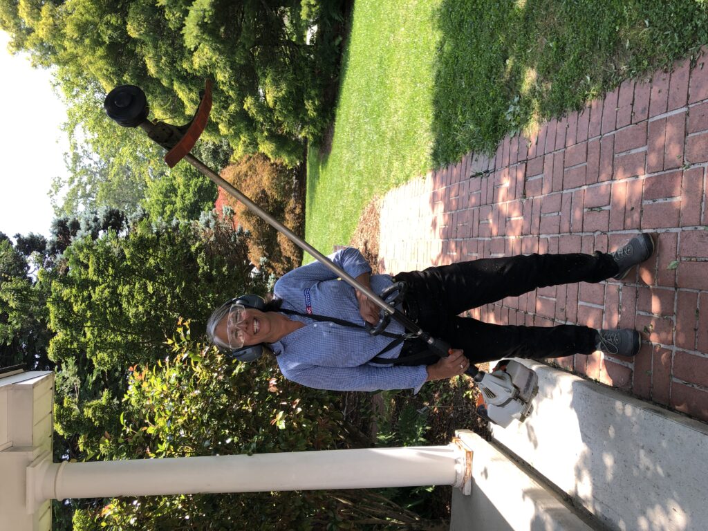 A smiling woman wearing a blue and white striped shirt, safety glasses, and ear protection stands on a brick path holding a weed trimmer.
