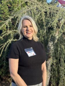 A smiling blonde woman wearing a black top and a name badge stands in front of a leafy tree.